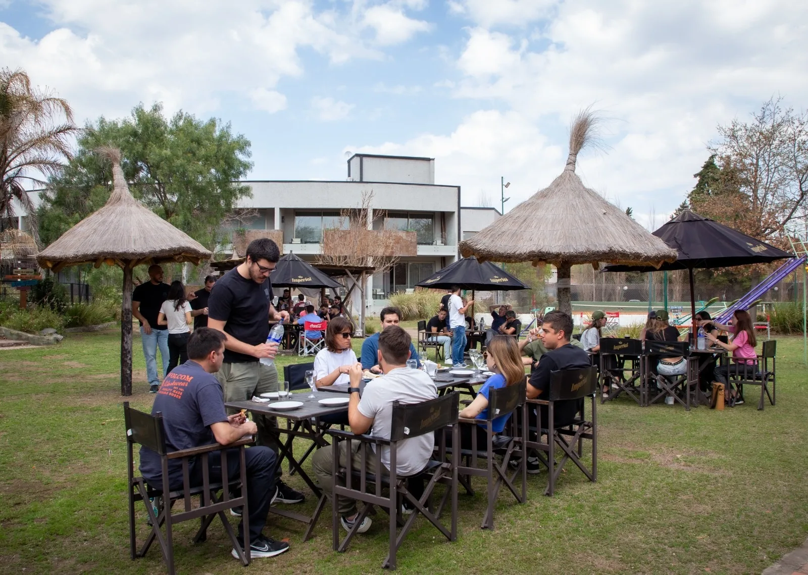 Grupo de una empresa almorzando en mesas al aire libre bajo sombrillas de paja, con el hotel al fondo
