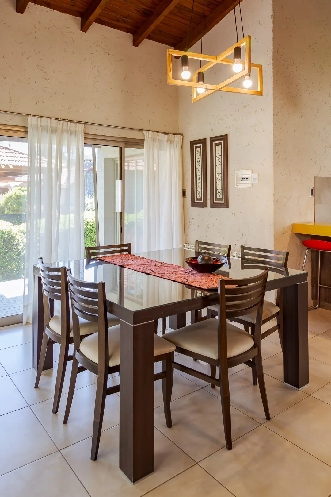 Comedor de una casa con mesa de vidrio oscuro, seis sillas, techo de madera y ventanal con cortinas