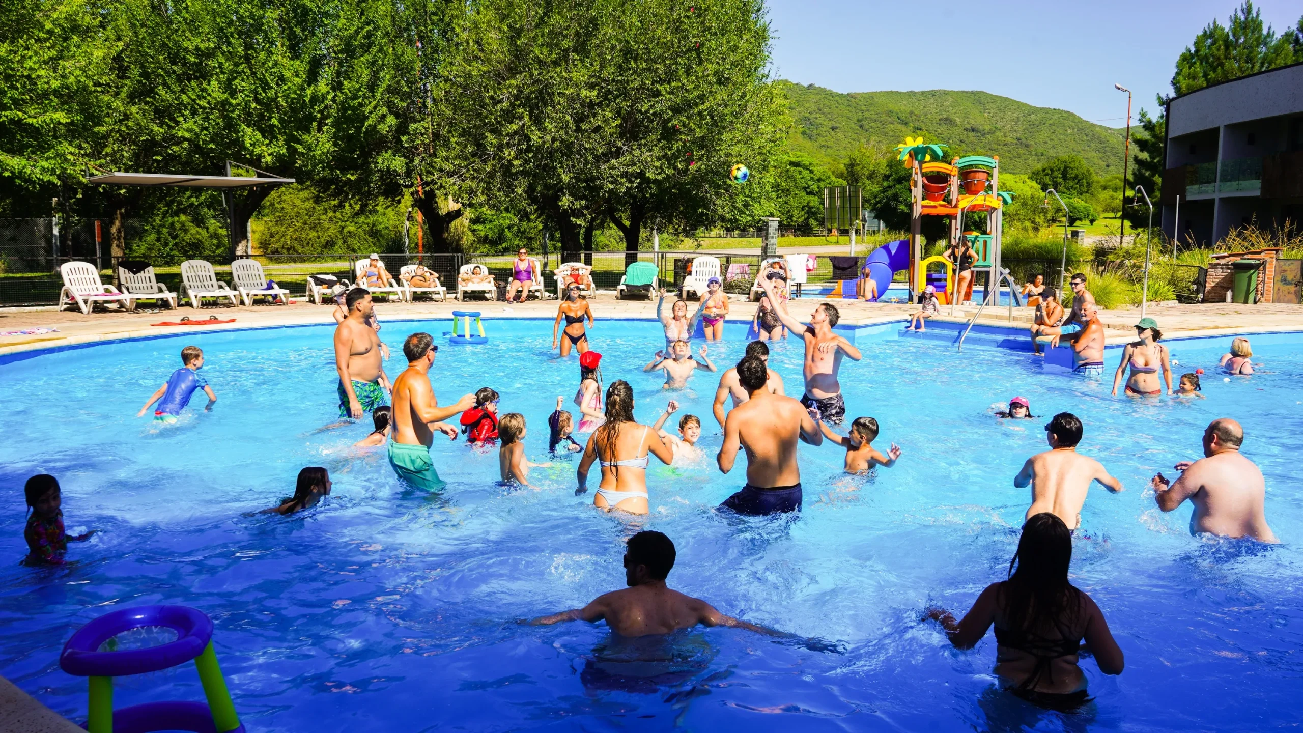 Grupo de adultos y chicos jugando con aros y pelotas en la pileta principal, con el hotel terminado y las sierras al fondo