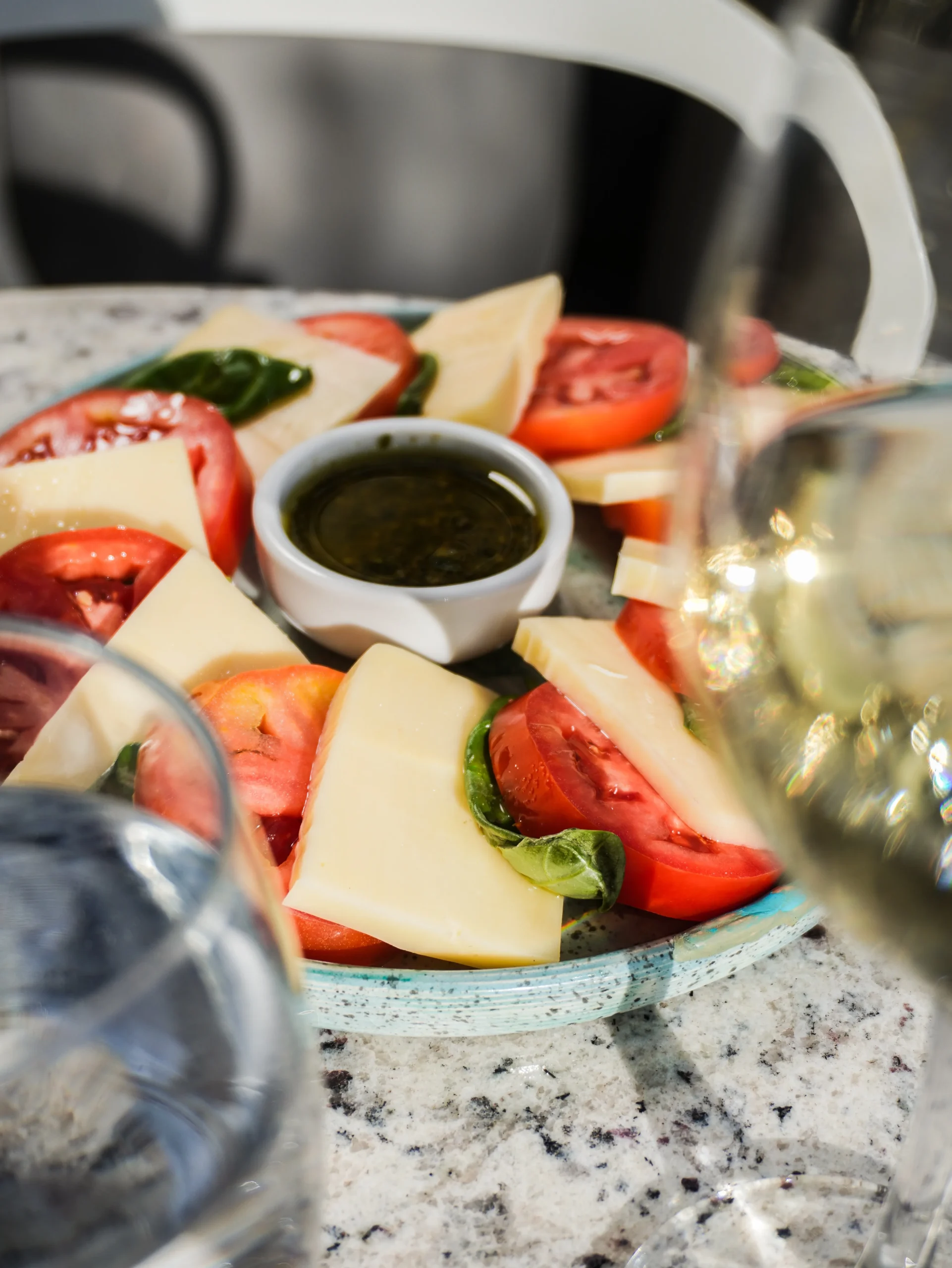 Plato de tomate, queso y albahaca con aderezo, junto a copas de vino sobre una mesa de granito