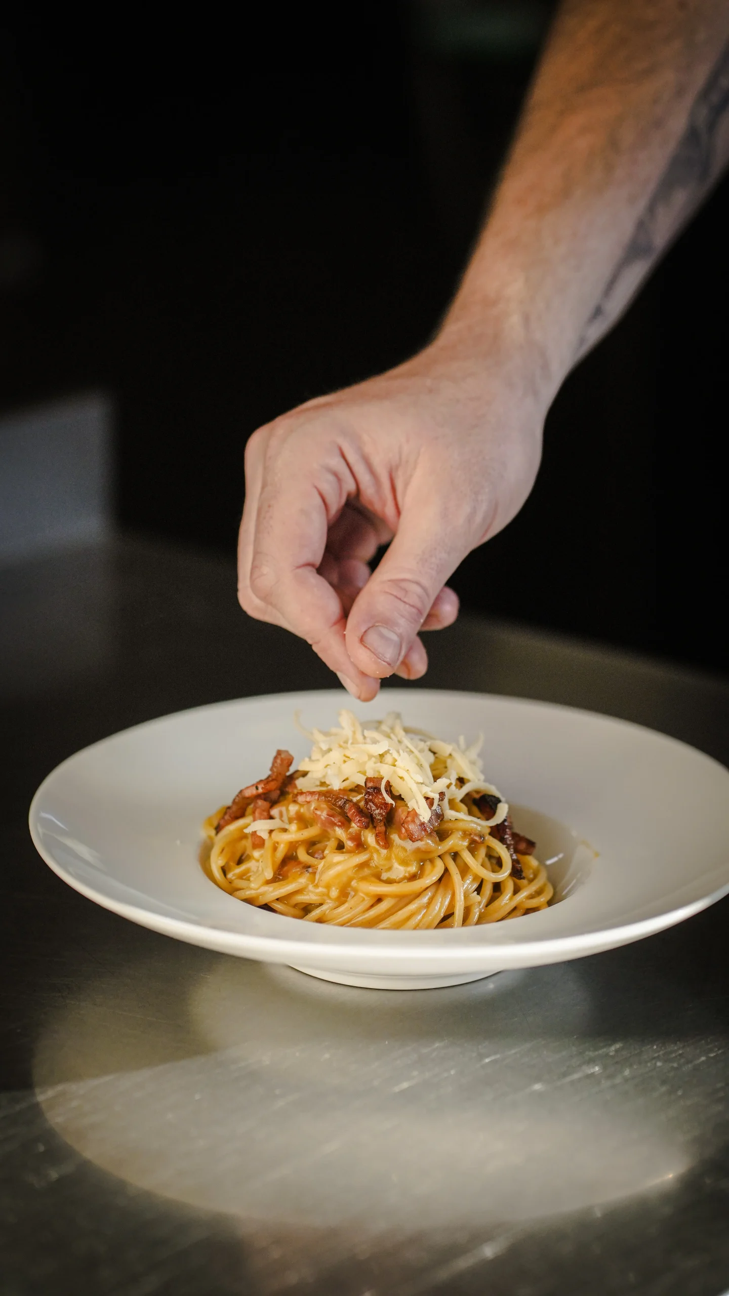 Mano del chef espolvoreando queso sobre un plato de spaghetti, sobre una mesada de acero