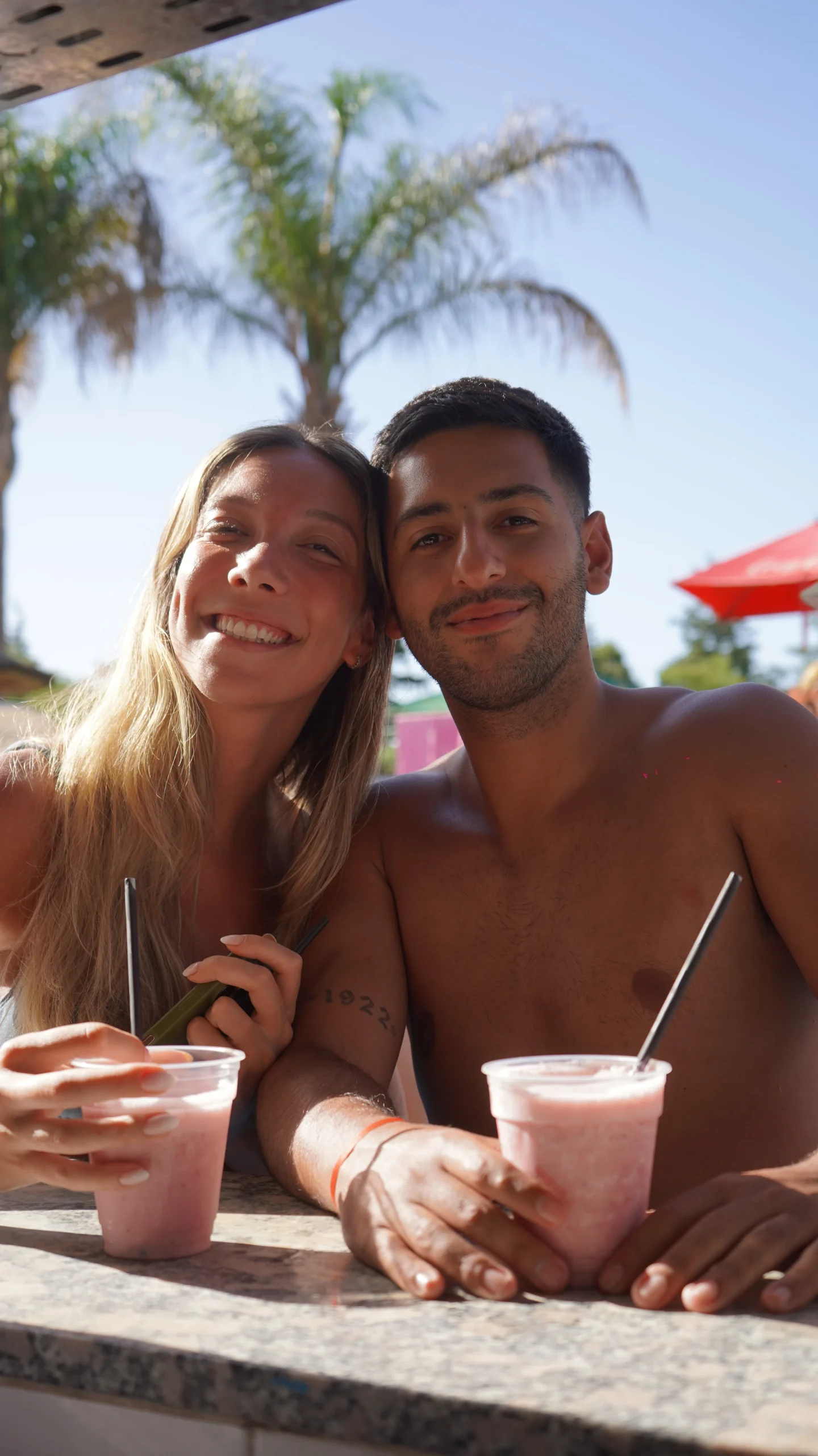 Pareja sonriente con licuados de frutilla apoyados en la barra del bar de la pileta, con palmeras detrás