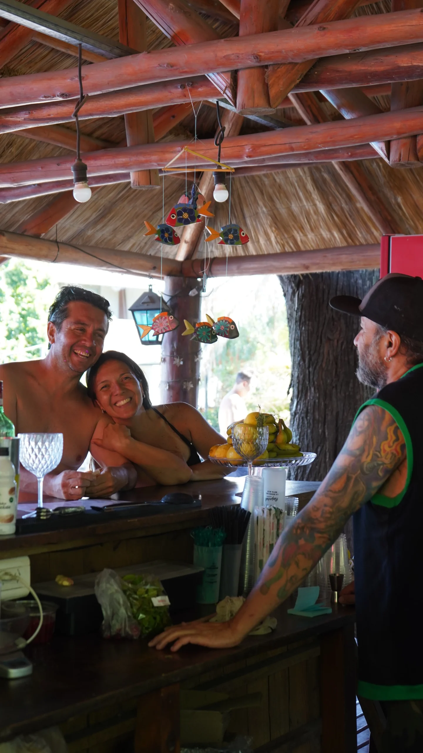 Pareja sonriendo en la barra del bar Santinhos, con techo de paja y troncos, atendida por el barman