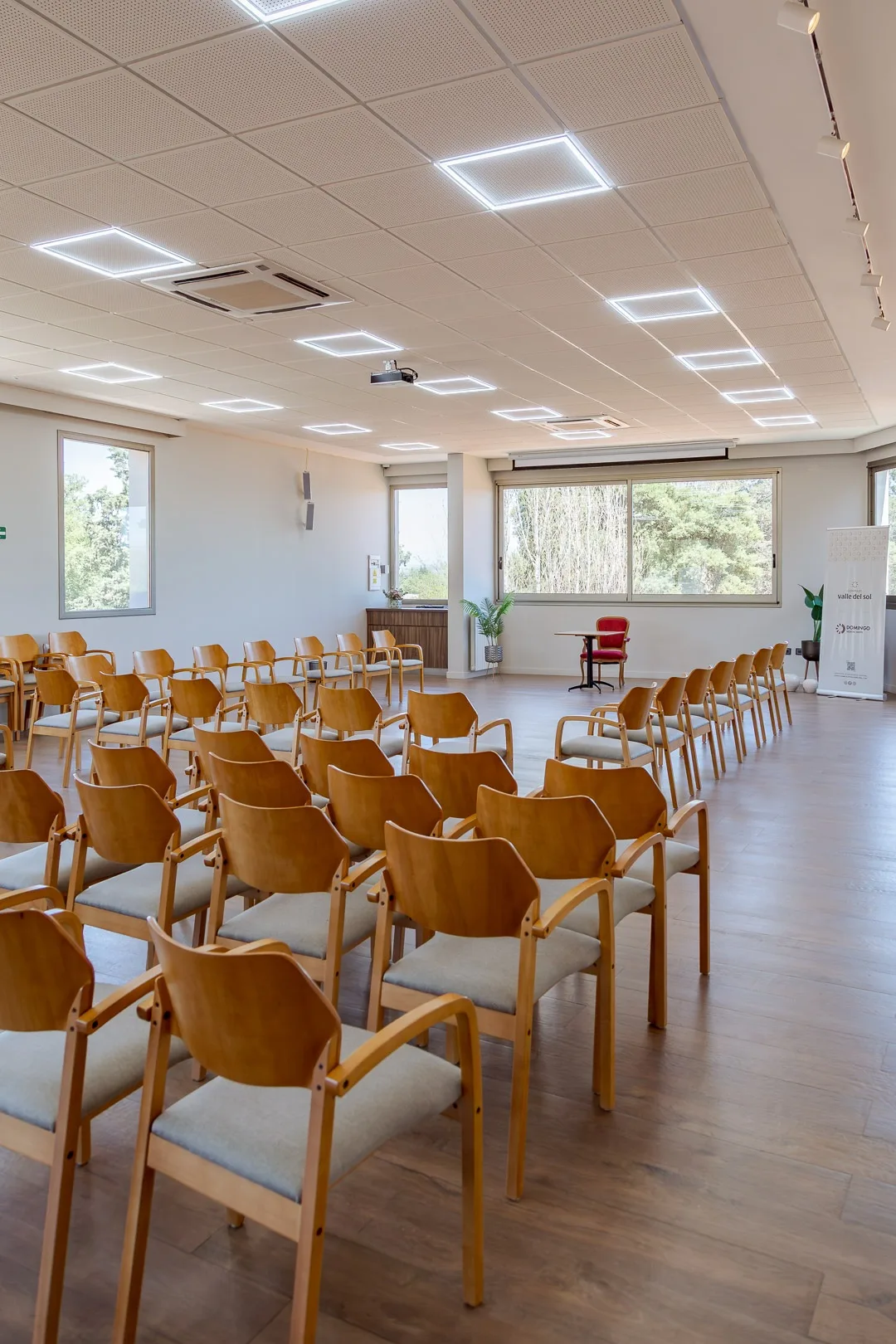 Salón de convenciones con sillas de madera en filas tipo auditorio y ventanales
