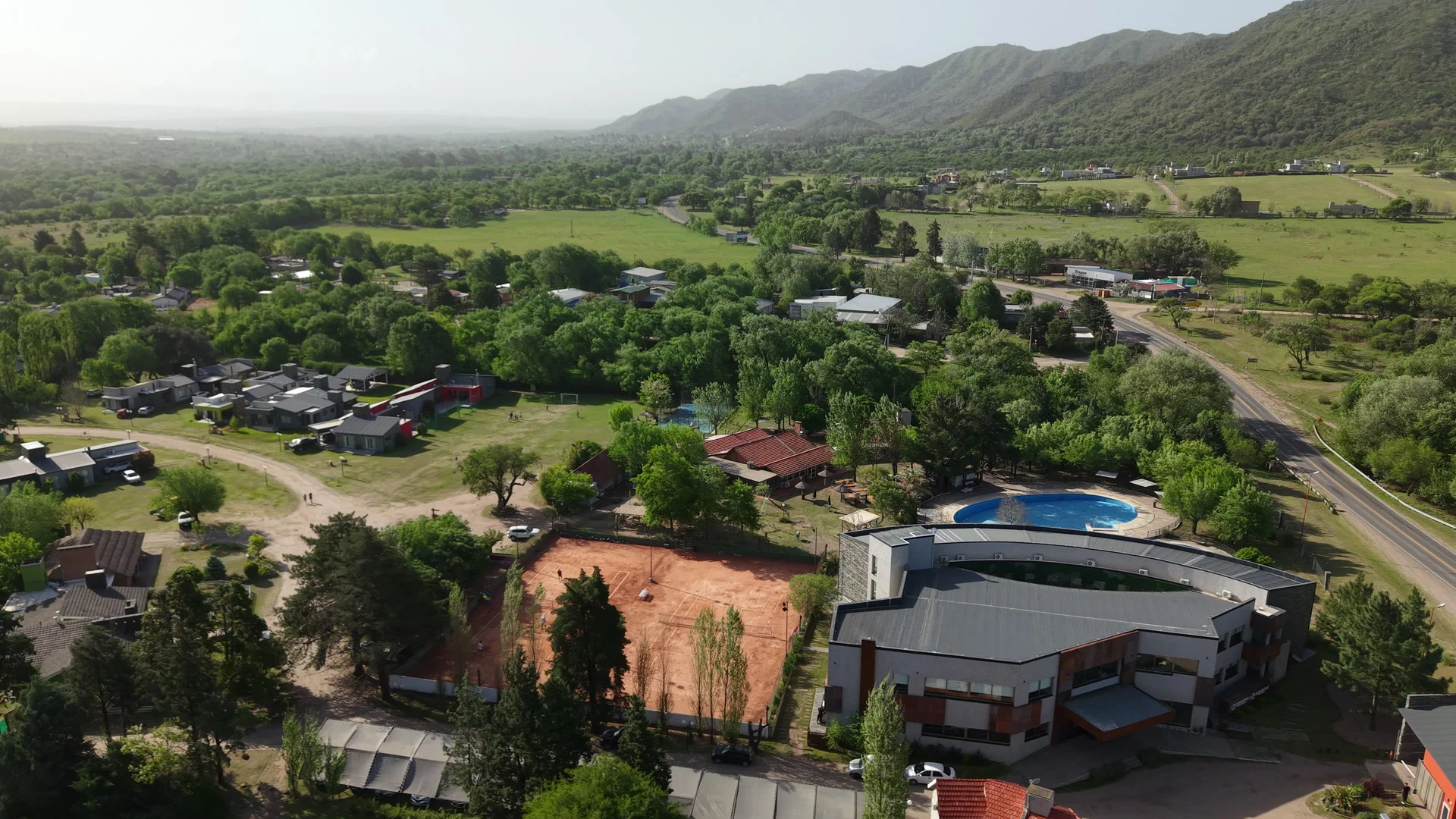 Vista aérea del complejo Valle del Sol con el Domingo Design Hotel, la cancha de tenis y la pileta, rodeados de arboleda y con las sierras al fondo
