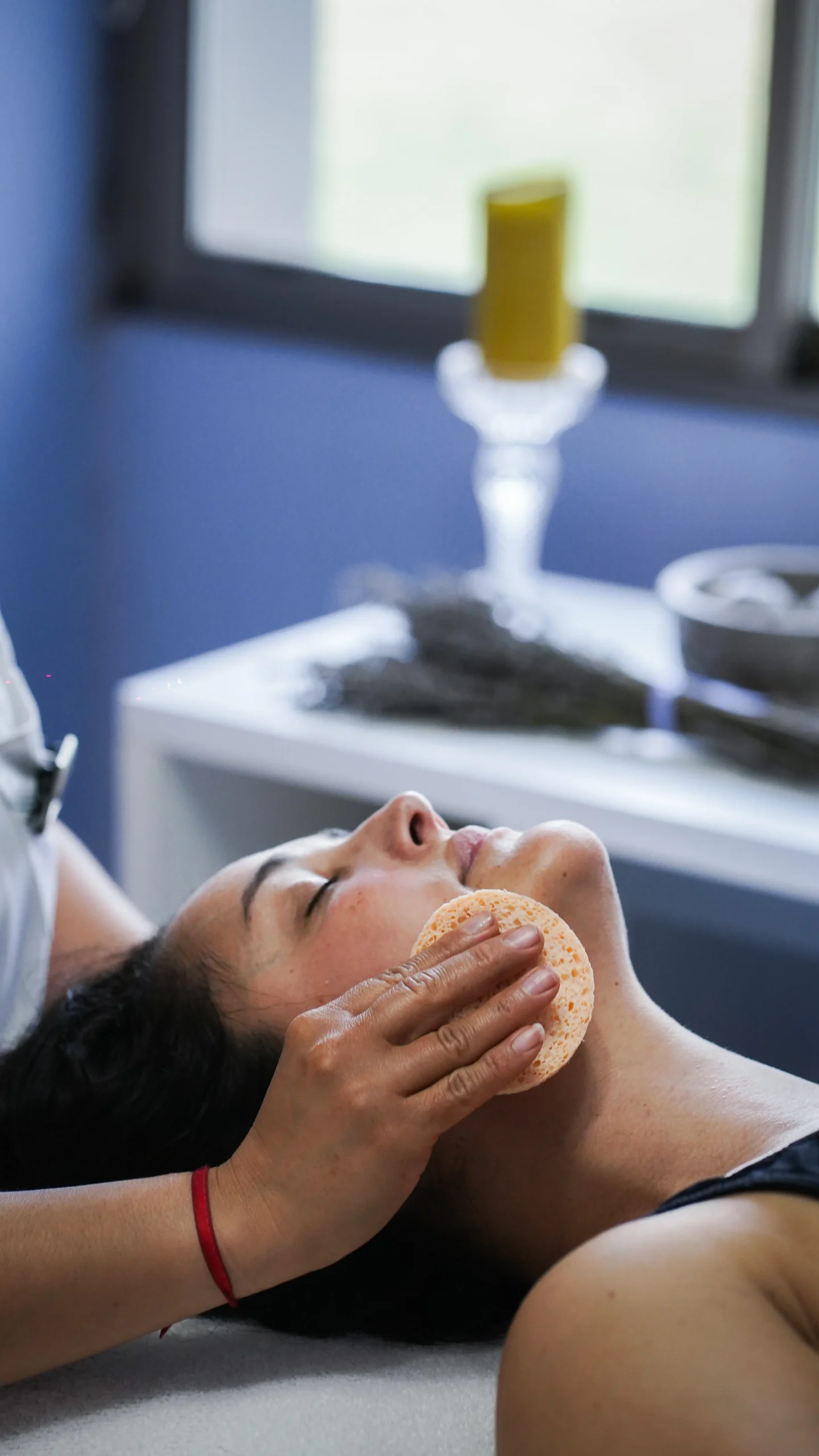Terapeuta limpiando el rostro de una mujer recostada en la camilla del spa, con una vela y lavanda detrás