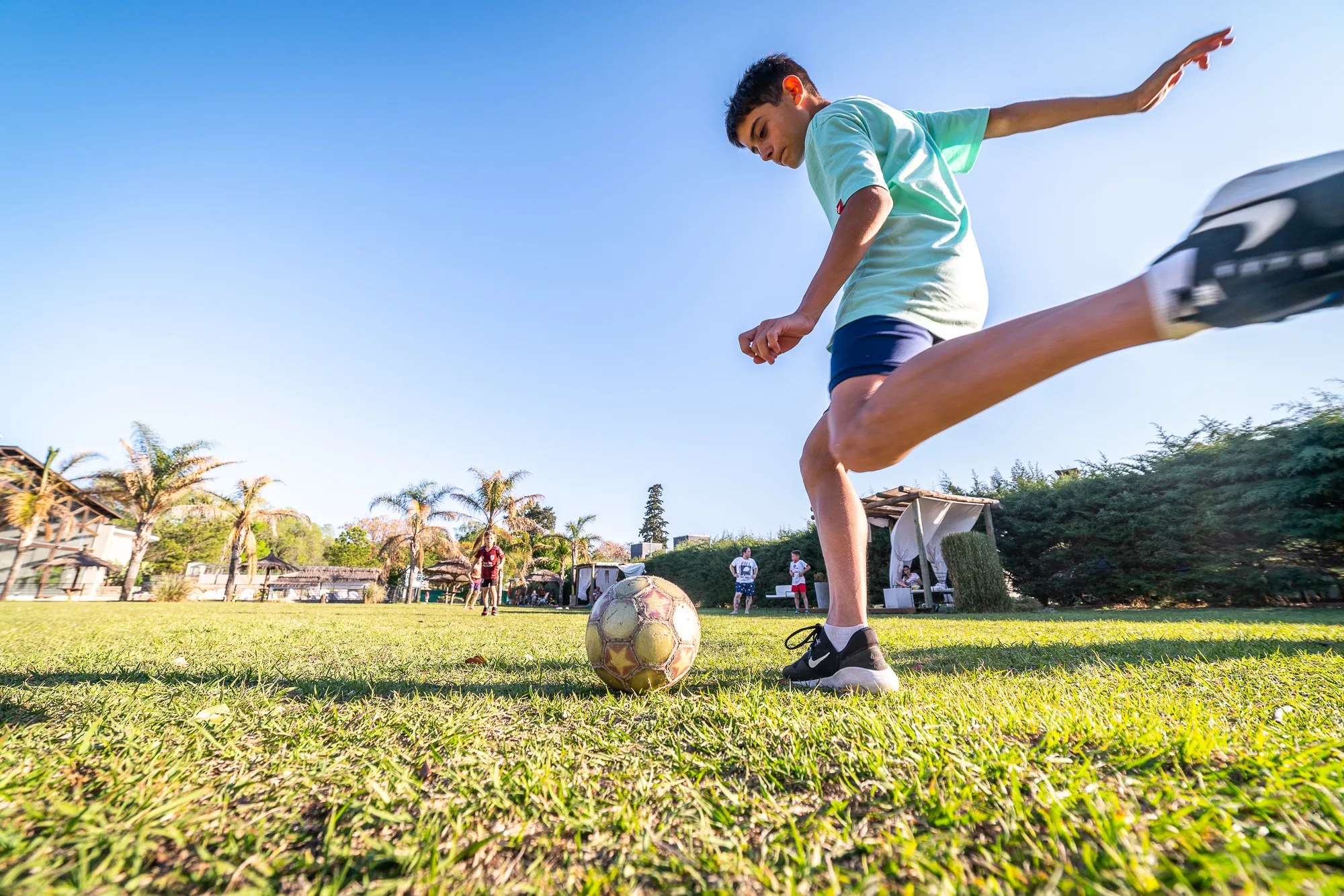 Chico pateando una pelota de fútbol en el césped, con palmeras y gente al fondo