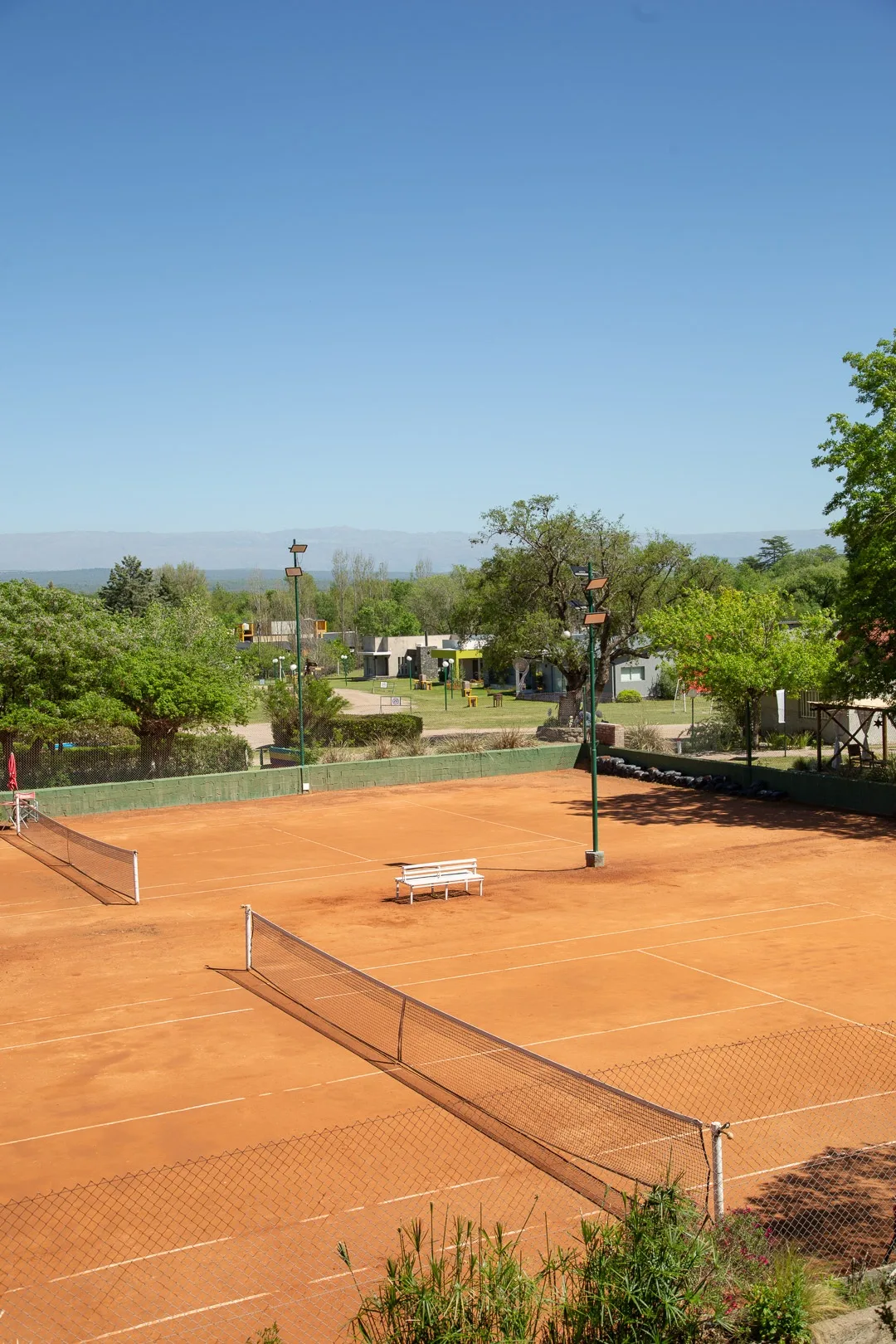 Cancha de tenis de polvo de ladrillo vista desde arriba, rodeada de árboles y con las sierras al fondo