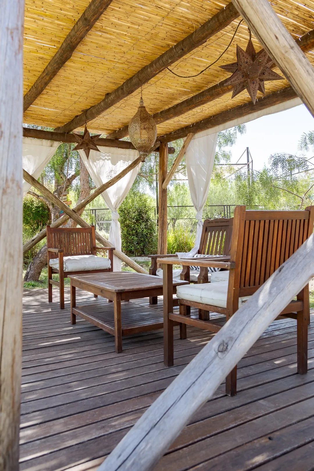 Interior de un gazebo de madera con techo de cañas, sillones con almohadones y cortinas blancas