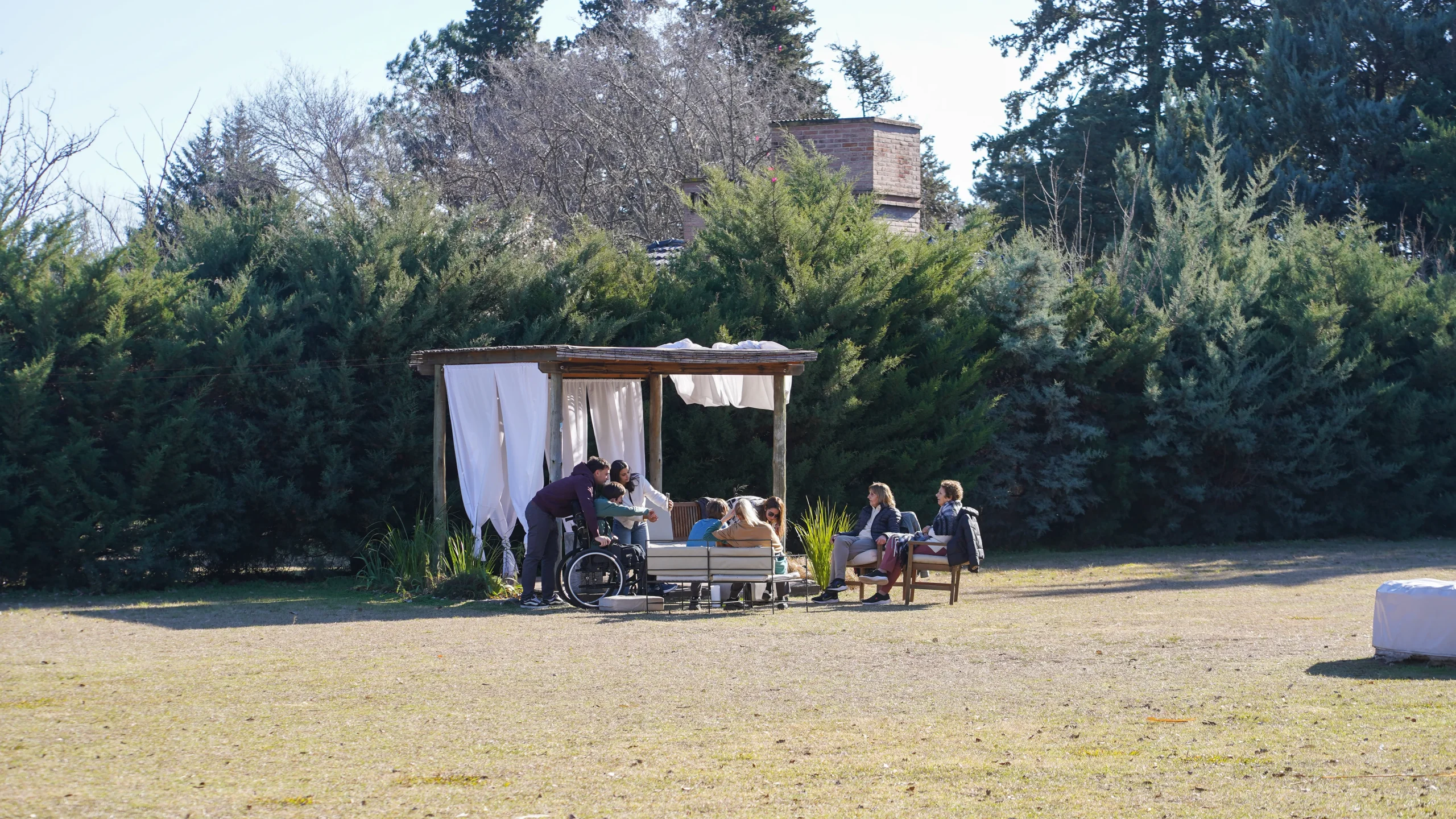 Grupo de personas reunido en un gazebo con cortinas blancas en el parque, rodeado de cipreses