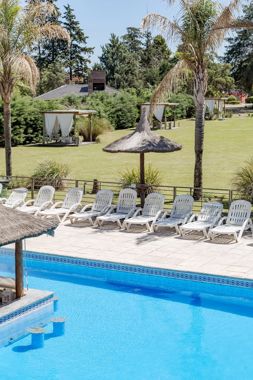 Pileta exterior con reposeras blancas y sombrilla de paja, y detrás el parque con gazebos de cortinas blancas